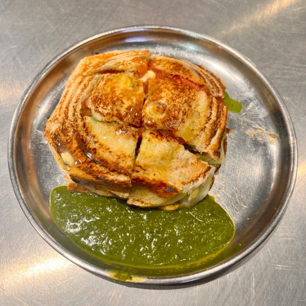 A steel plate on which sits a veg toast sandwich cut into 6 pieces, and a side of green chutney.